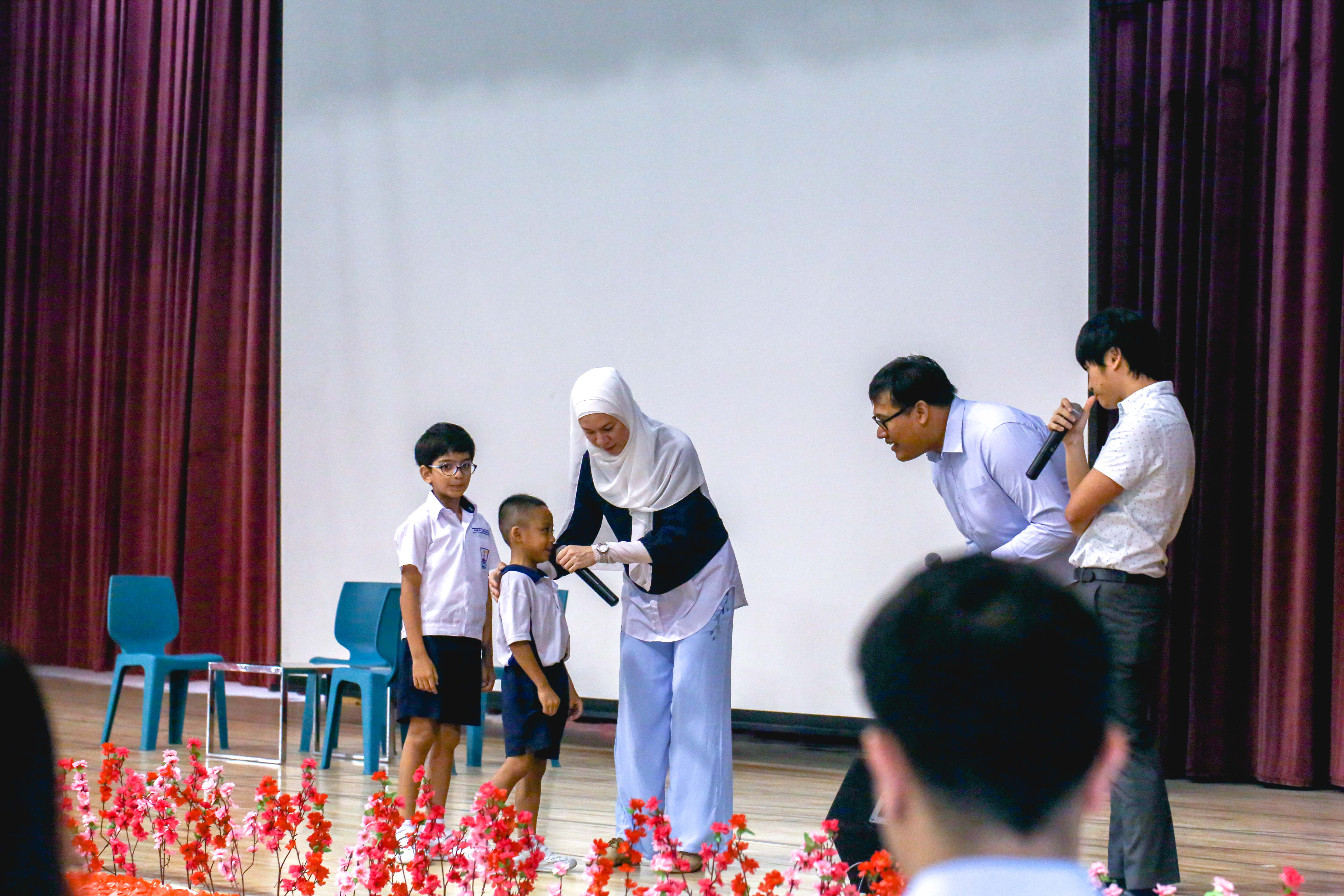 Two adults with microphones lean toward young students in uniforms on a stage decorated with red flowers.